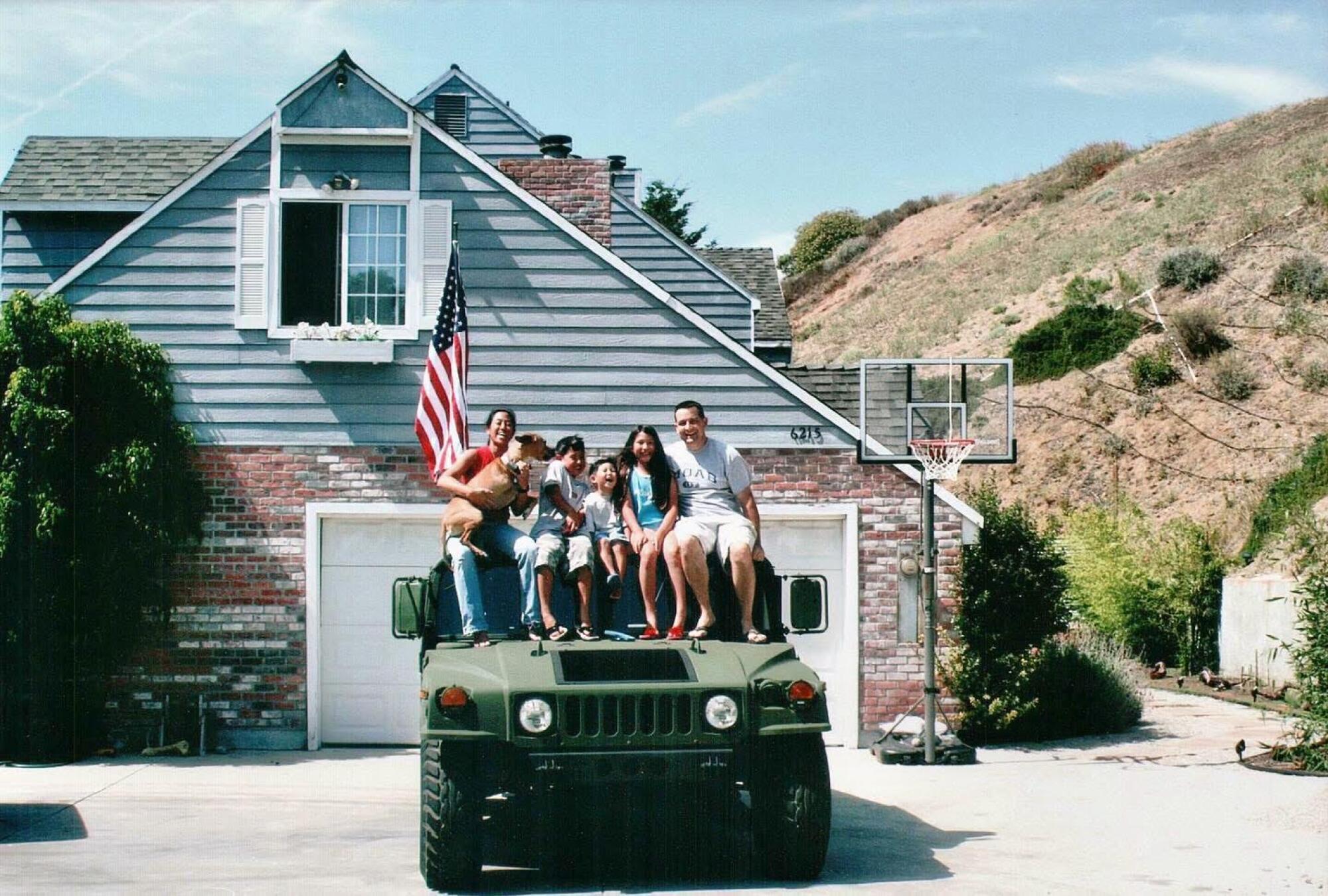 A family sits on top of a car, in front of a house with an American flag.