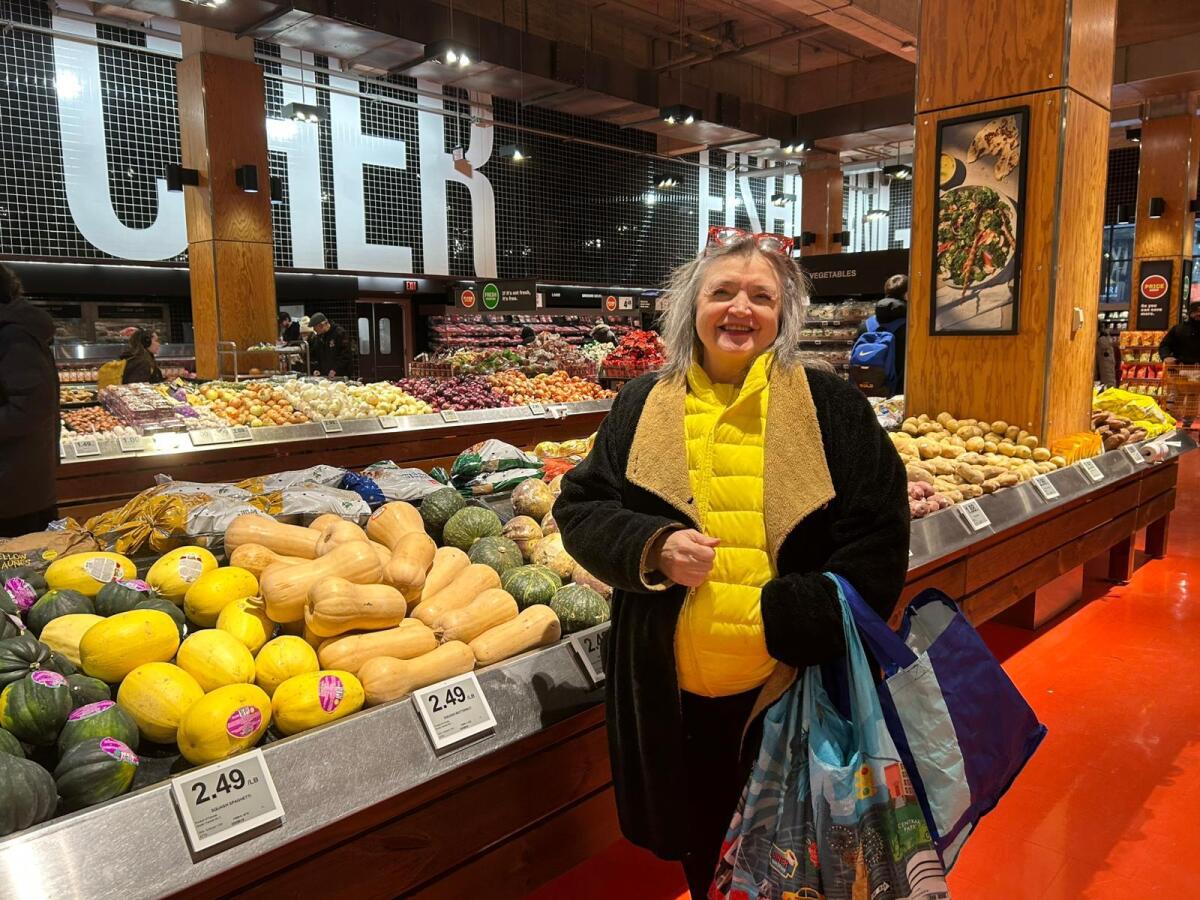 A woman in a dark jacket and yellow vest, holding shopping bags, stands near a counter with produce