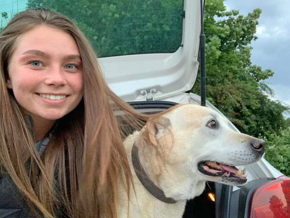 A young woman with long brown hair smiles while sitting with her light-colored dog in the back of a car.