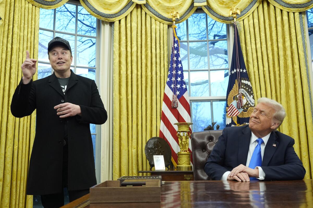 A man in a dark suit and blue tie, seated at a desk, smiles and looks toward a man in dark clothes, pointing up as he speaks