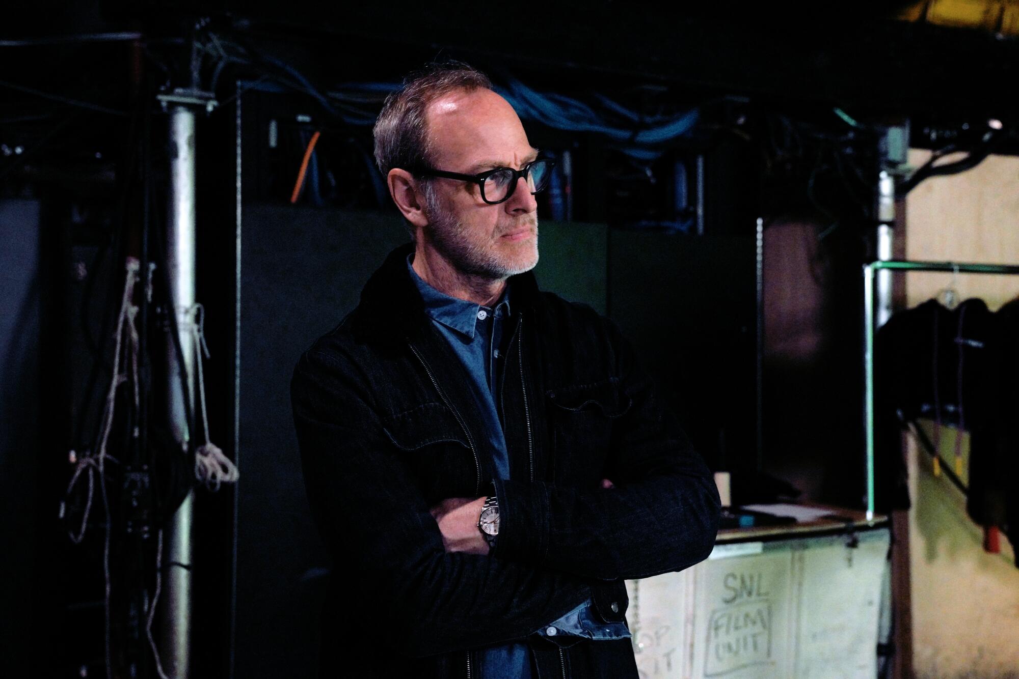 A man wearing black-rimmed glasses and a dark blazer stands with his arms folded inside a studio.