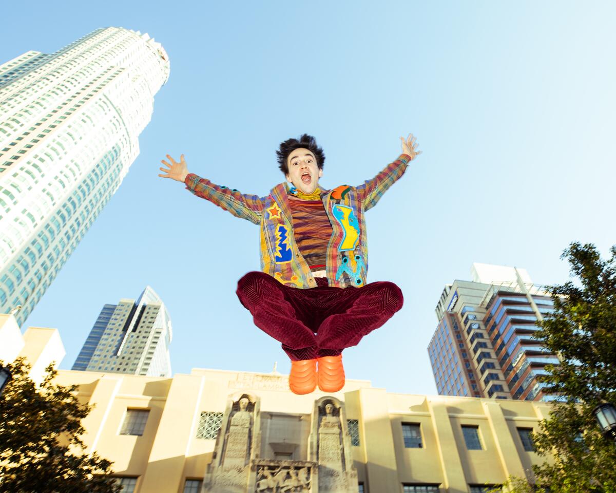 Jacob Collier, wearing bright clothing, jumps in the air in front of the L.A. skyline.