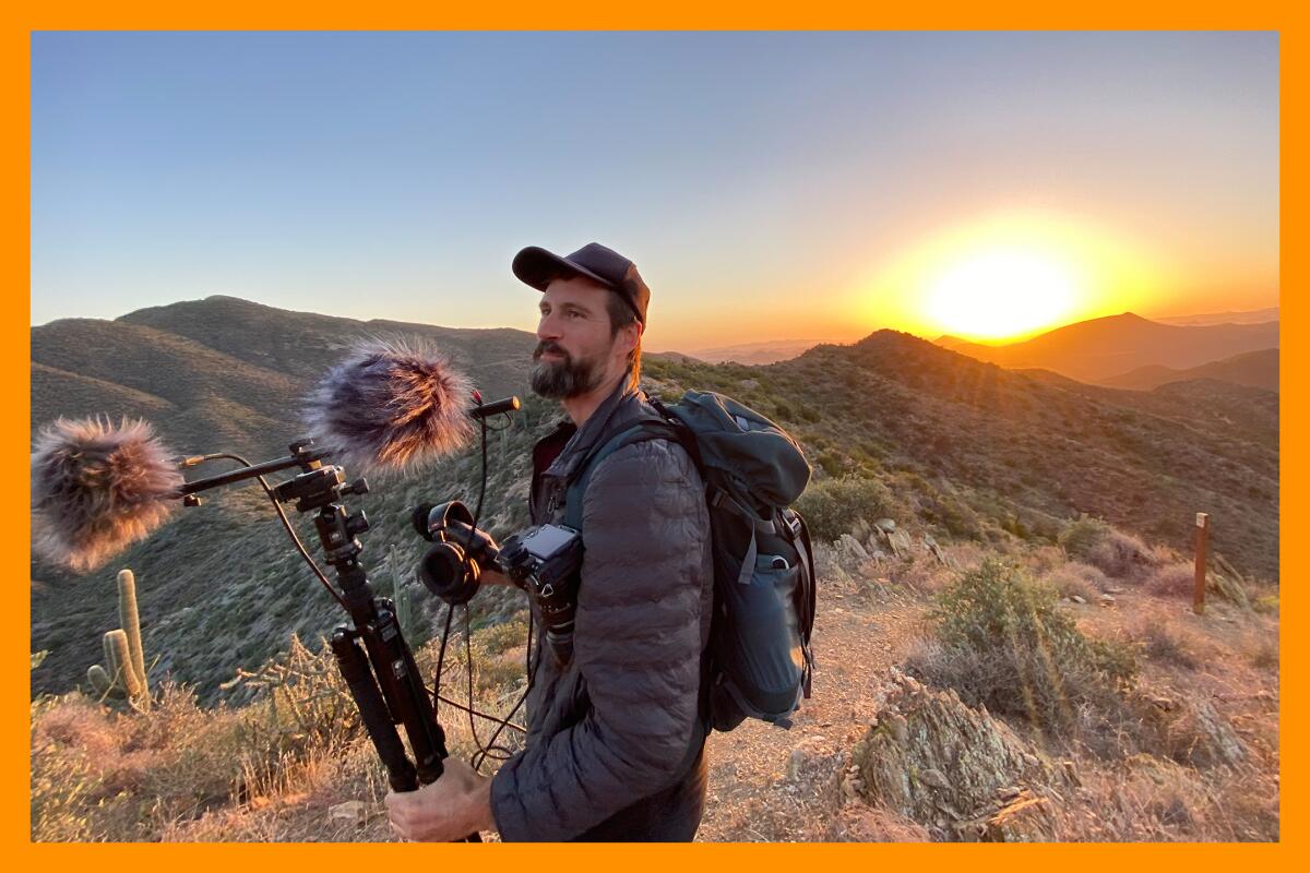 A person stands on the top of a mountain with audio recording equipment. The sun sets in the background.