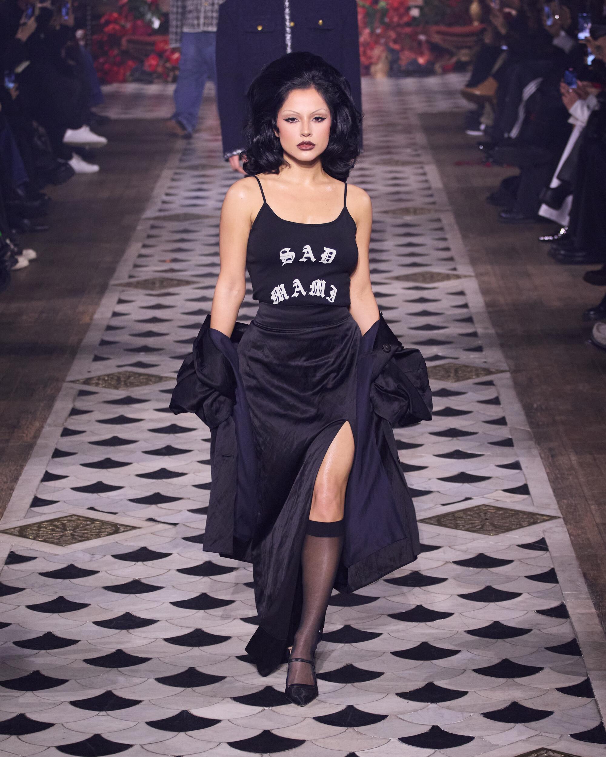 Becky G in a black ensemble walks on a black-and-white patterned floor at a fashion show