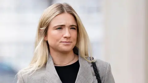 PA Media Ms Mewis is wearing a grey blazer, black top and has a gold necklace on. She has blonde hair and is wearing small hoop earrings. She has a straight face and looking beyond the camera. 