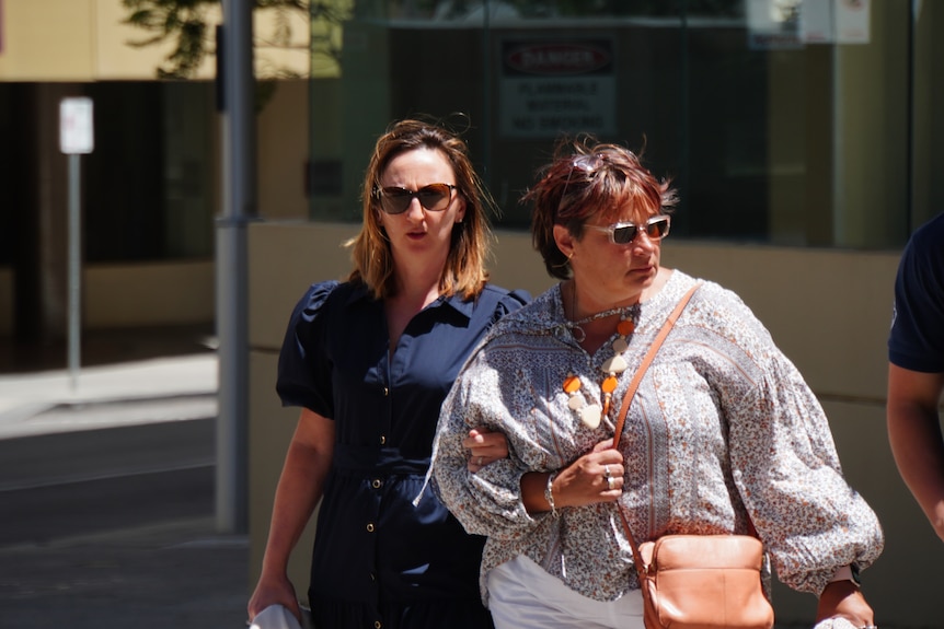 A woman clutches the arm of another woman as she arrives at court, both women wearing sunglasses