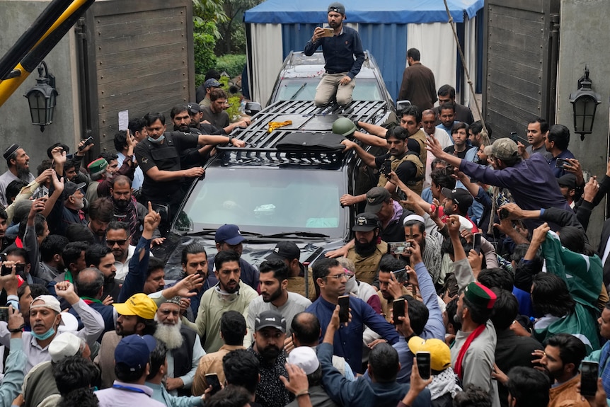 People crowd around vehicle carrying Imran Khan out of gates of residence.