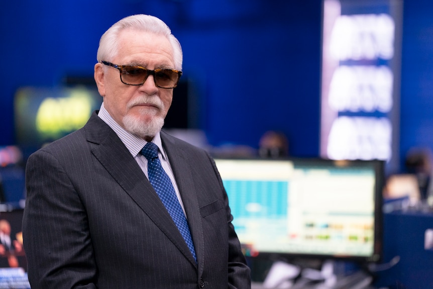 A production still of Logan in wearing sunglasses in the newsroom.