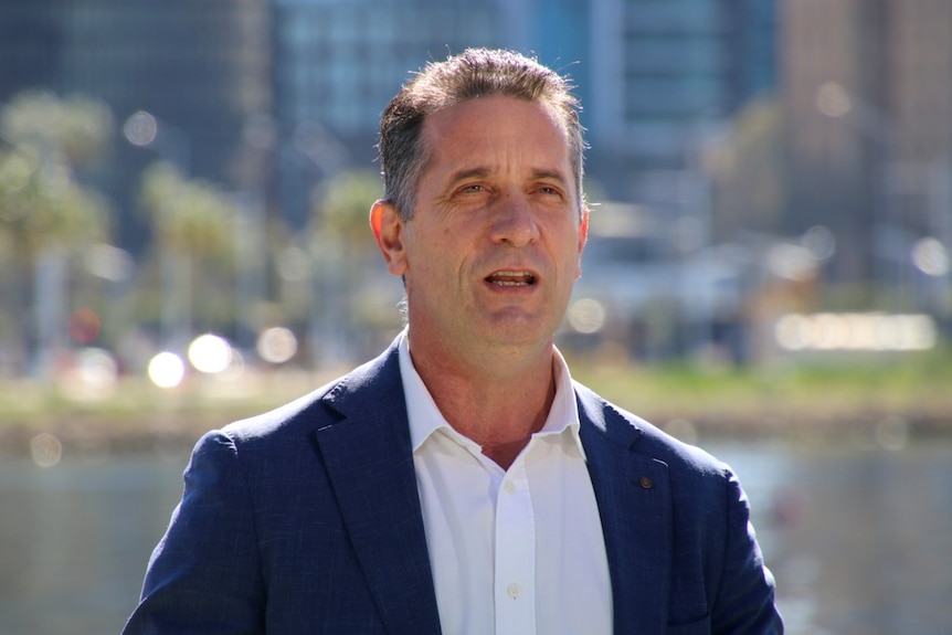 A man wearing a navy blazer and white shirt stands on a deck with the Perth CBD in the background.