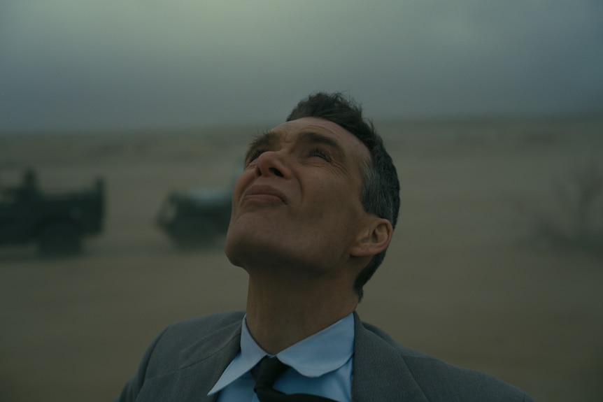 A white man with light brown hair and wearing a grey 40s suit, stands in a windswept and gloomy desert landscape.