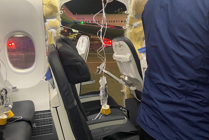 Oxygen masks dangle in front of several plane seats with the window missing from a plane.