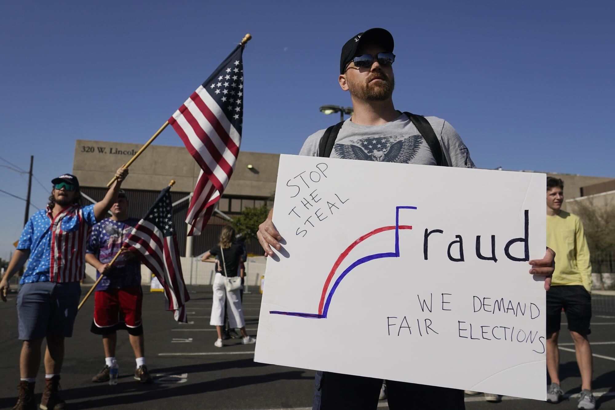 A man among a small group holds a sign that says, "Fraud - Stop the Steal - We Demand Fair Elections"