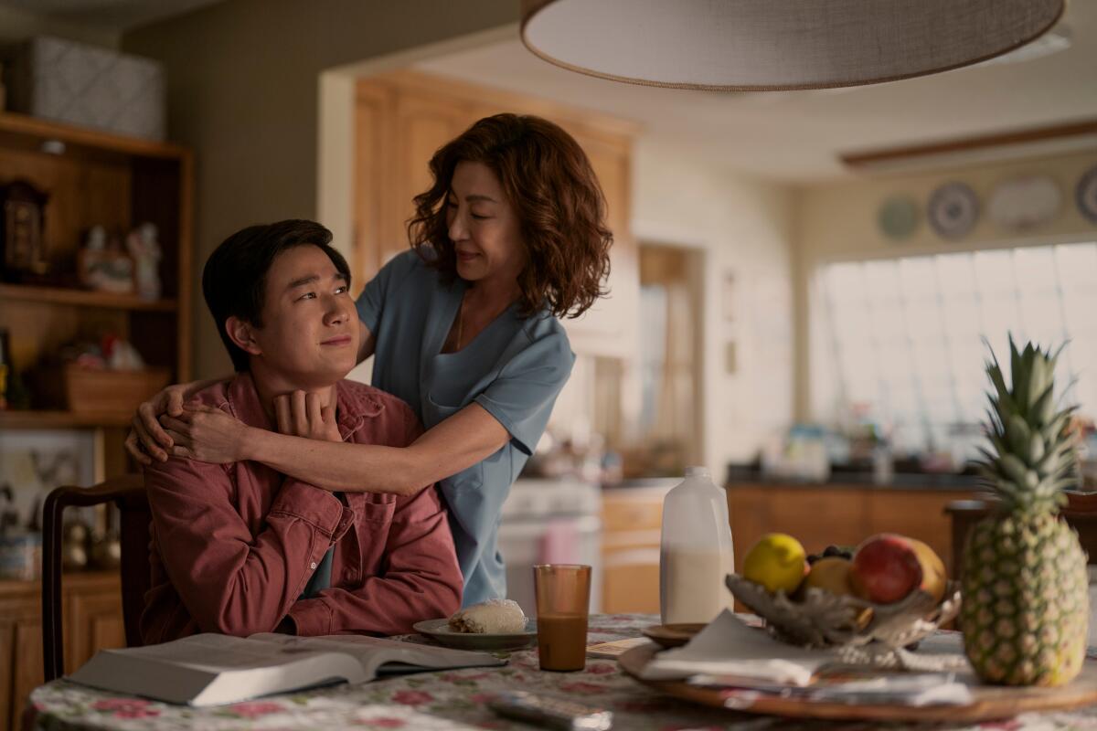 A man sits at a kitchen table as his smiling mother wraps her arms around his shoulders.