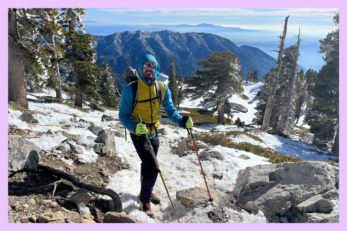 A  man in winter gear and holding a walking stick in each hand stands on a snow-covered hiking trail