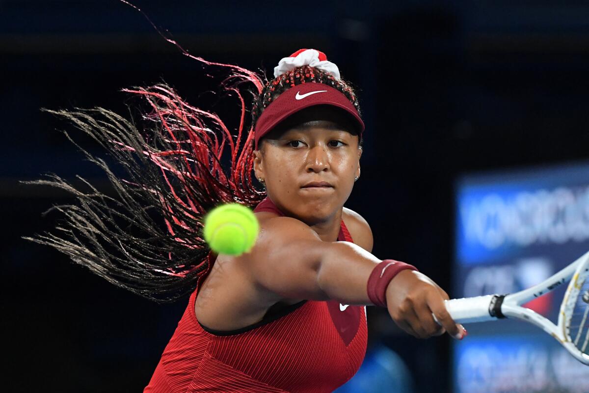 Noami Osaka in a red outfit, hair flying behind her, smacks a tennis ball toward the camera. 