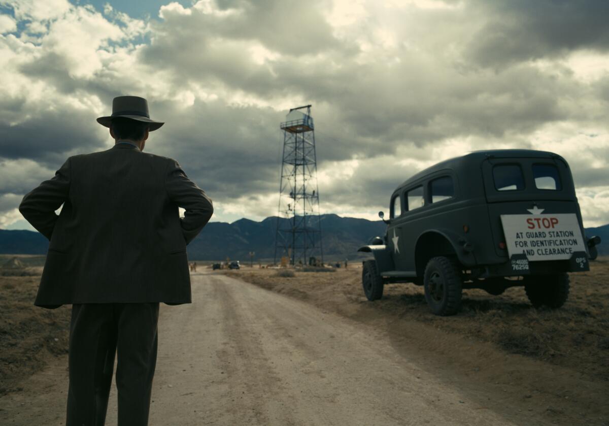 A man in suit and hat stands on a dirt road under a cloudy sky looking into the distance.