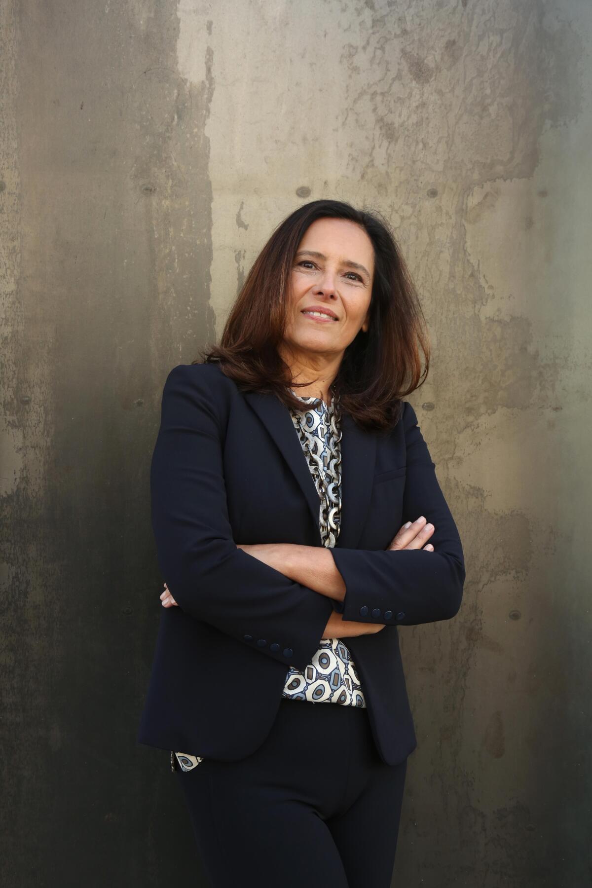 A woman in a dark blazer with her arms crossed in front of a wall.