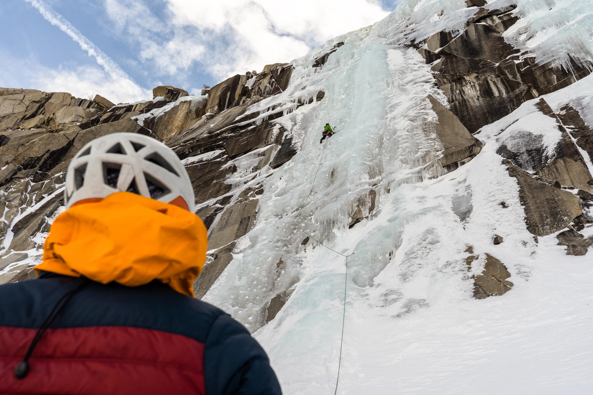 A man wearing a helmet looks at a frozen waterfall hugging a cliff. 