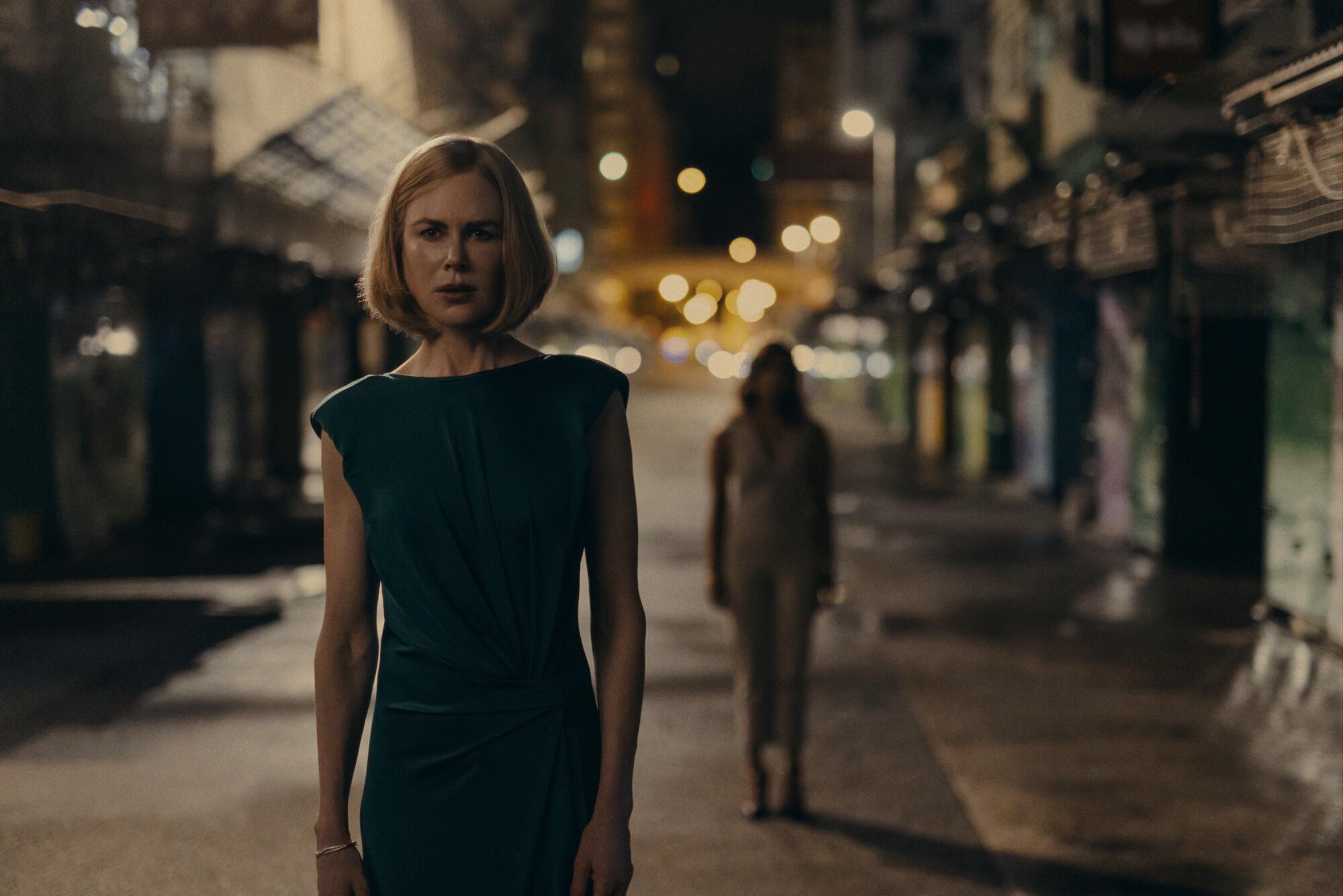 A woman in black dress stands in a dark alley as a person walks behind her.