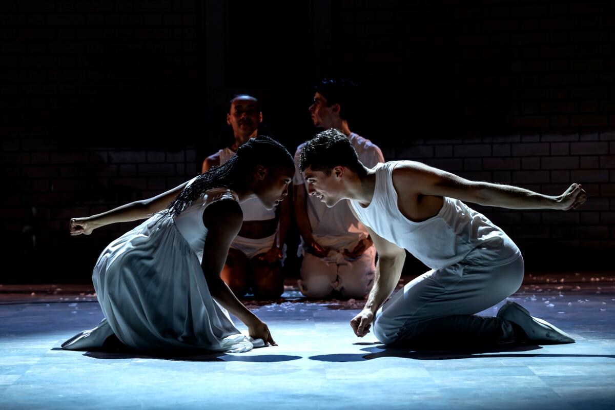 Two dancers wearing white kneel and bow before each other, their heads almost touching.