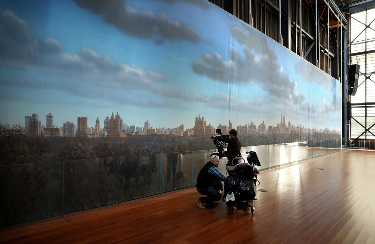 A video crew is shown filming a painted backdrop of a city skyline inside a large, wood-floored warehouse buildilng.