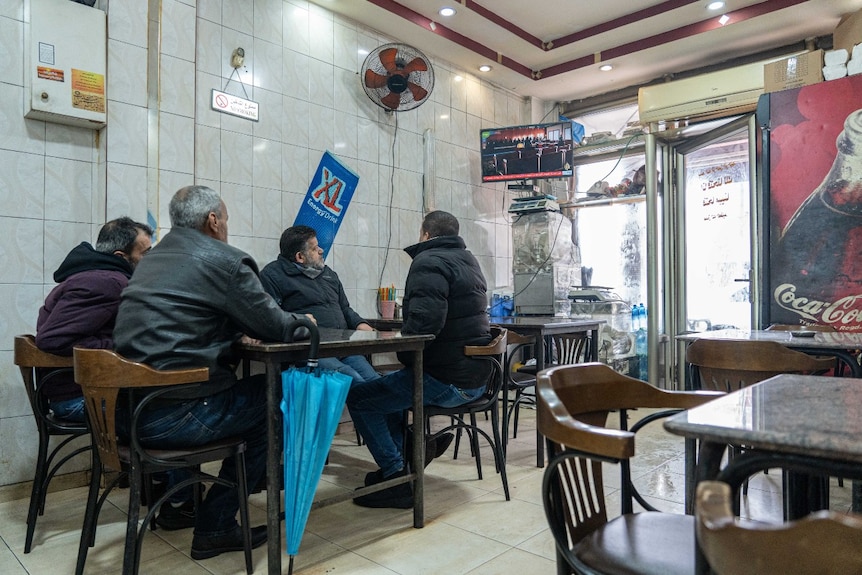 Four men sitting at a table, in a restaurant, watching a news broadcast on a television