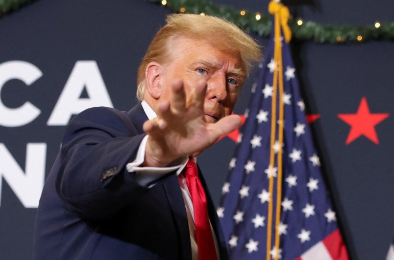 Donald Trump extends his hand, his fingers outstretched, toward the crowd at a December 2023 campaign rally. Behind him is a US flag and a campaign banner.