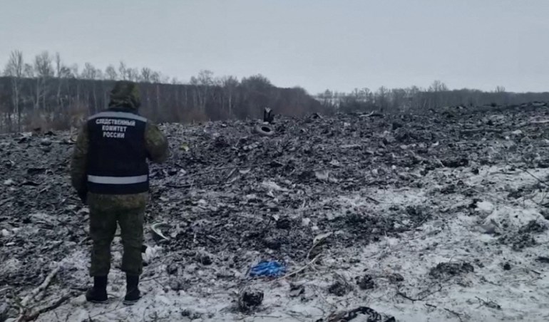 Black and grey debris scattered in the snow after the crash of the Ilyushin-76 plane. An investigator is standing to one side, looking at the scene. 