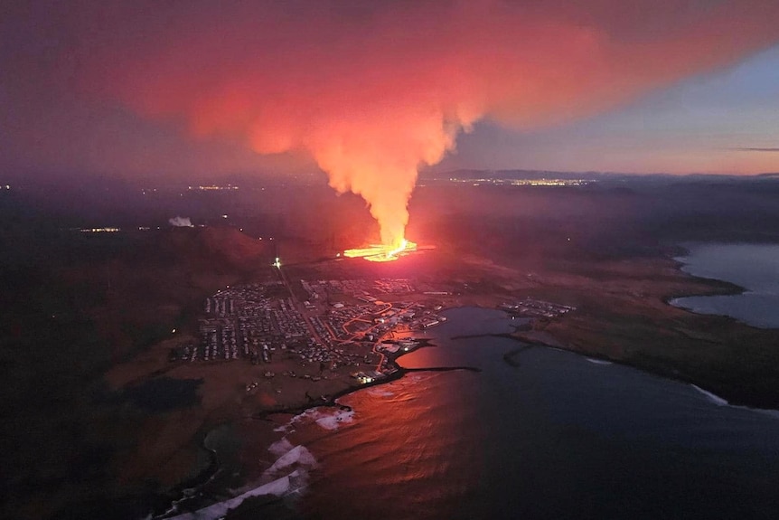 A long distance shot of orange smoke billowing into the dark night as a puddle of bright lava creeps towards a town