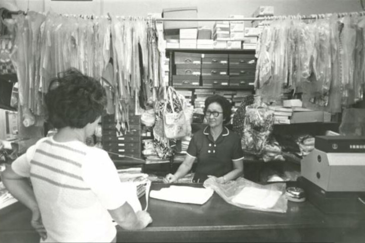 A black and white photo of a woman behind a counter with a book talking with another woman