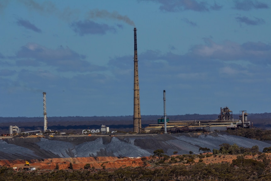 An industrial scene with a smoke billowing out of a tall smoke stack.   