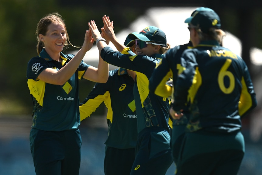 Five female cricketers for Australia converge in celebration, clapping hands, after a wicket