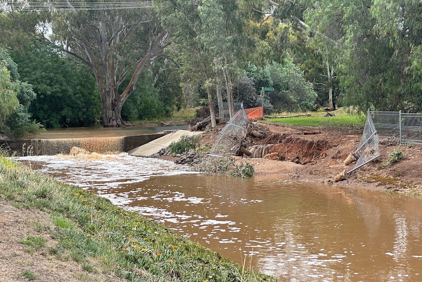 A flooded road spilling down into a creek, with a fallen fence lining the bank.