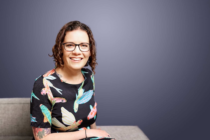 A woman smiles at the camera. She's sitting on a couch, wearing a dress with birds printed.