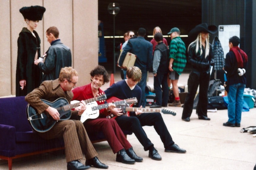 Three men in foreground playing guitar, with various people mingling in the background