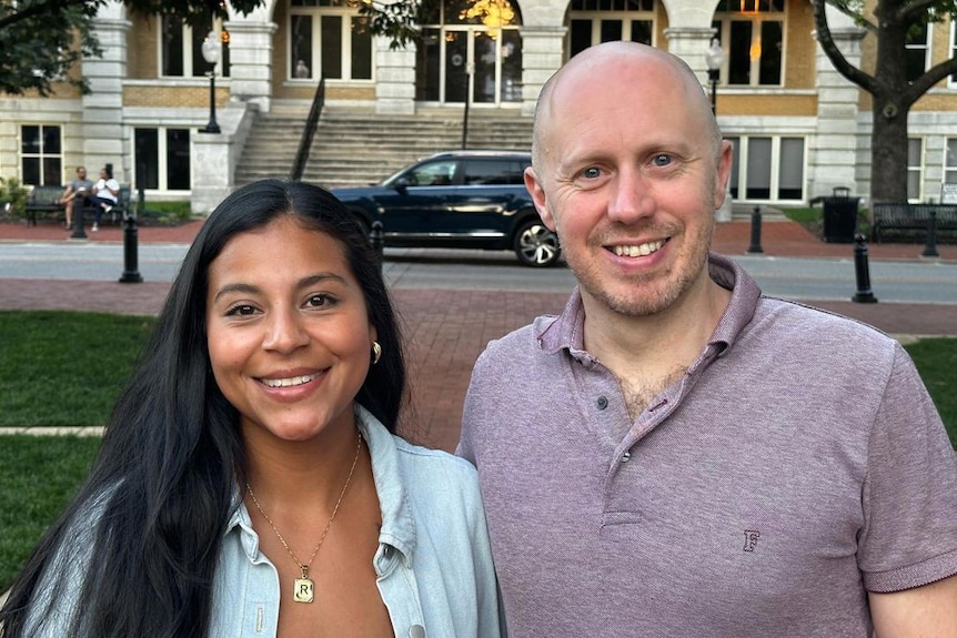 A man in a purple polo shirt poses for a photo next to a woman with dark hair who is wearing a blue shirt and necklace