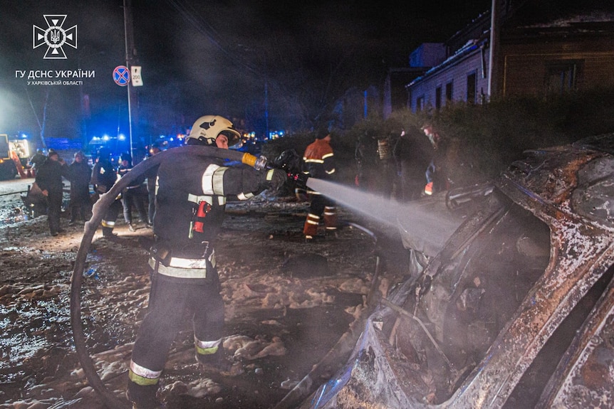 A person in long navy pants and a navy top with reflective white stripes holds a large hose as it sprays water.