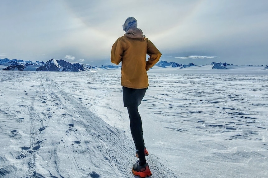 Woman in winter-wear runs on snow towards ice-capped hills with sun in front of her