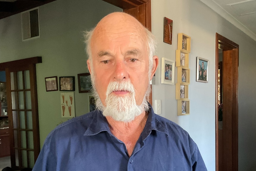 A light skinned man with short white hair and beard faces camera with disappointed expression, inside a house. 