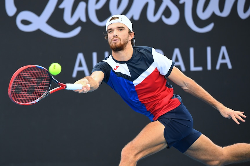 A man in a backwards cap holds onto a tennis racquet while trying to hit a tennis ball.