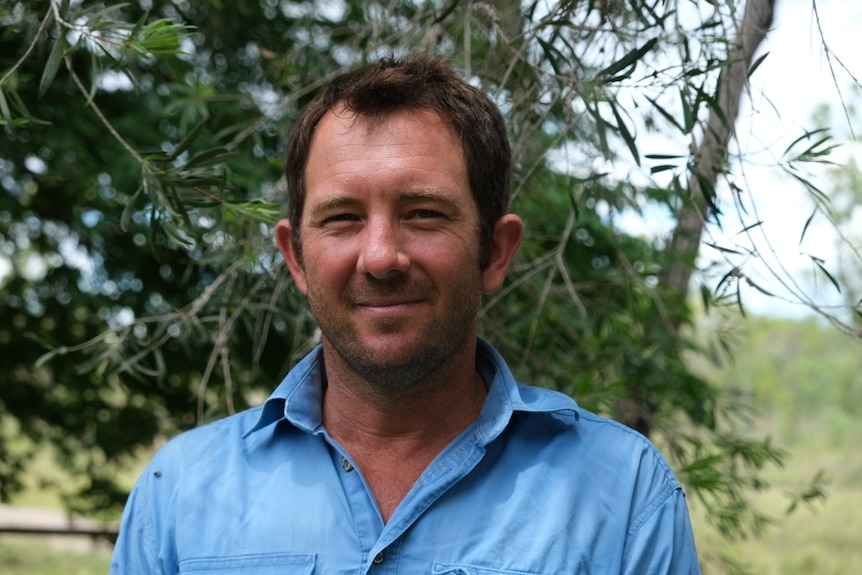 A man in a blue collared work shirt smiles standing in front of green leafy trees