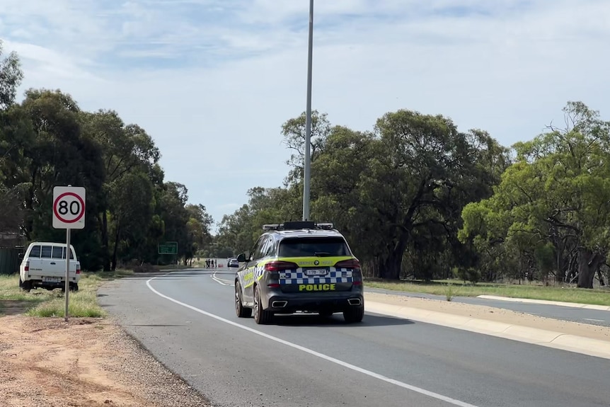 A police car is parked on a bend in the road beside an 80 kilometre an hour sign with detectives in the far background.