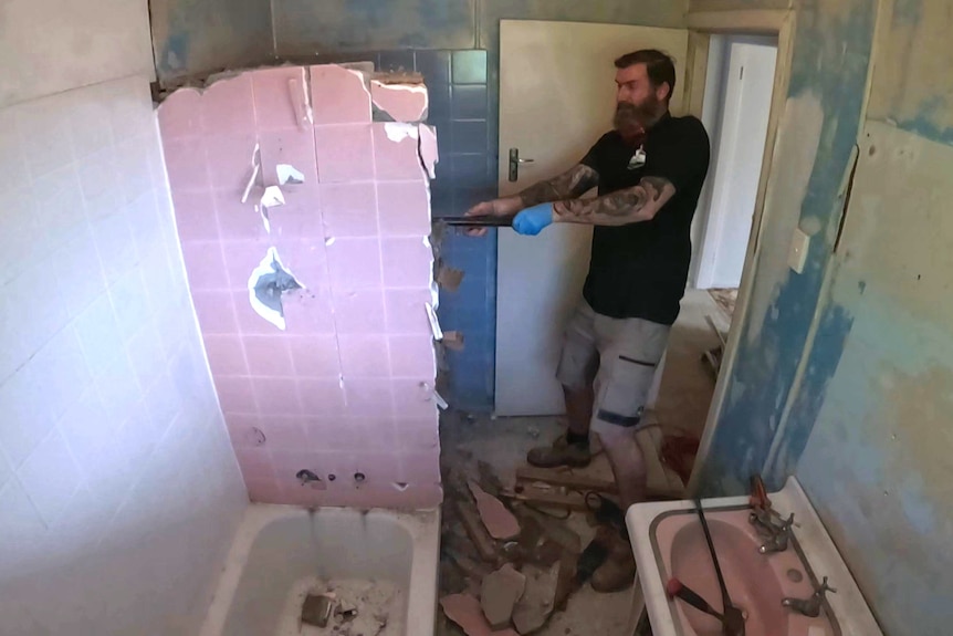 A wide angle shot of a man swinging a sledgehammer to get rid of a tiled wall in his bathroom 