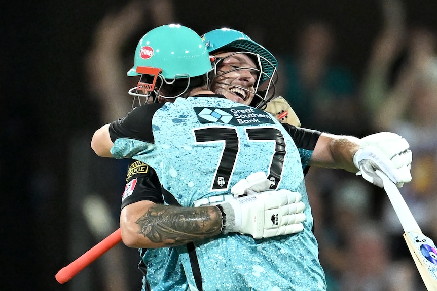 Two male cricketers, batting in the middle, hug each other in celebration.