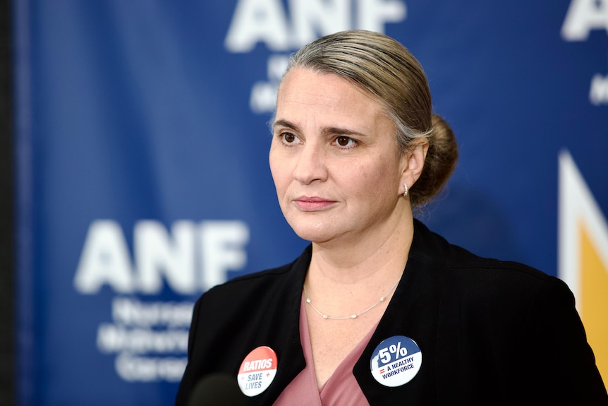 A middle-aged woman with a black blazer and a silver necklace. Her hair is in a bun and there's a medium blue ANF backdrop.