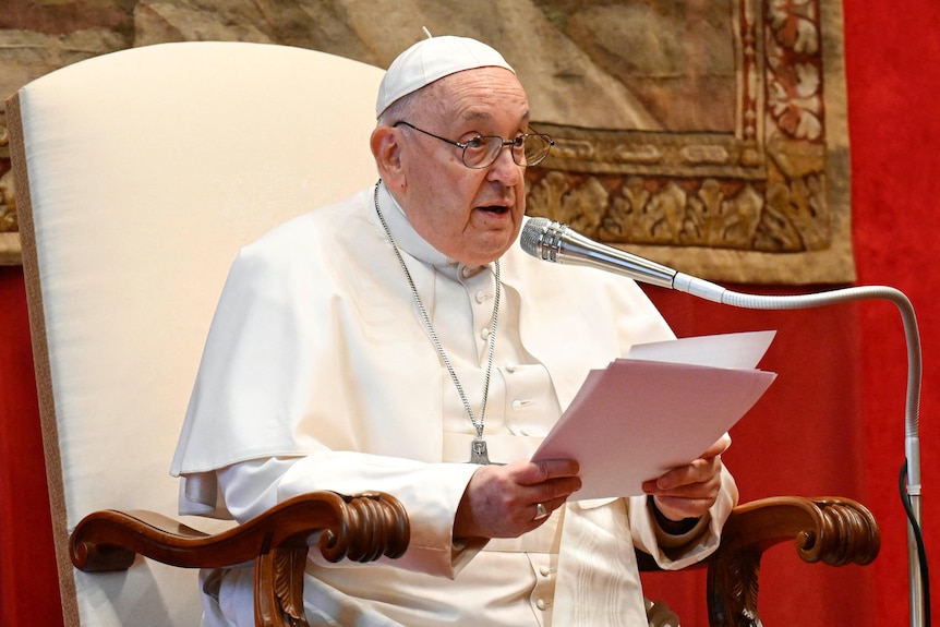 An elderly man in a white cap and gown reads from a piece of paper into a microphone.