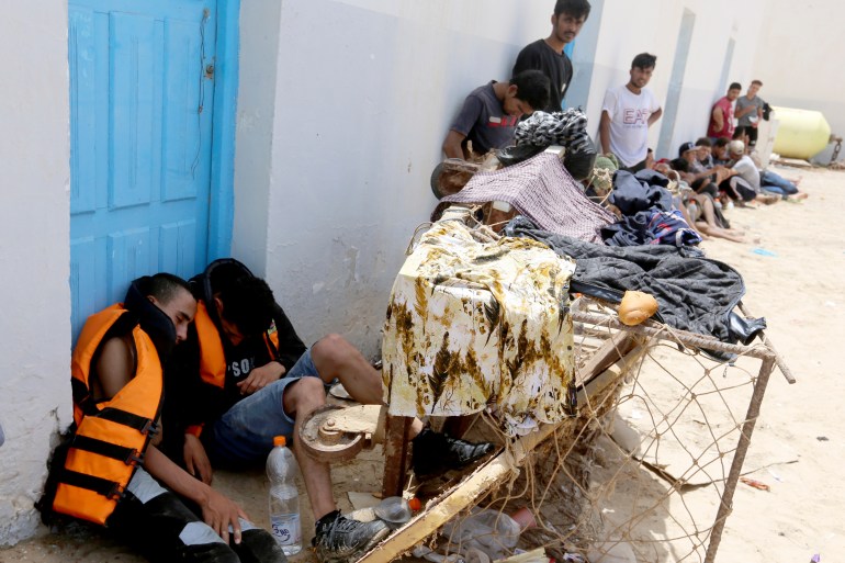 Migrants and asylum seekers, some of whom still wear their orange life vests, sit slumped on a street curb in the city of Ben Gardane. Behind them is a white wall with a blue door.