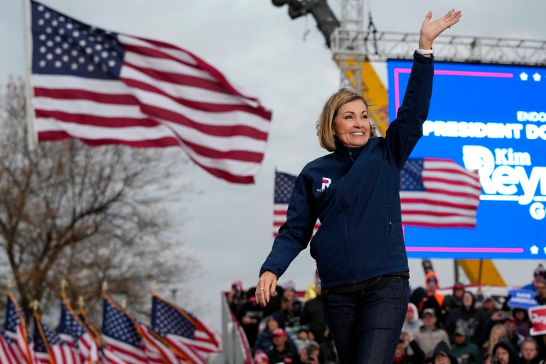 Kim Reynolds waves as she walks across a stage at a Trump rally, where two large American flags wave in the background. A blue electronic screen can also be seen behind her.