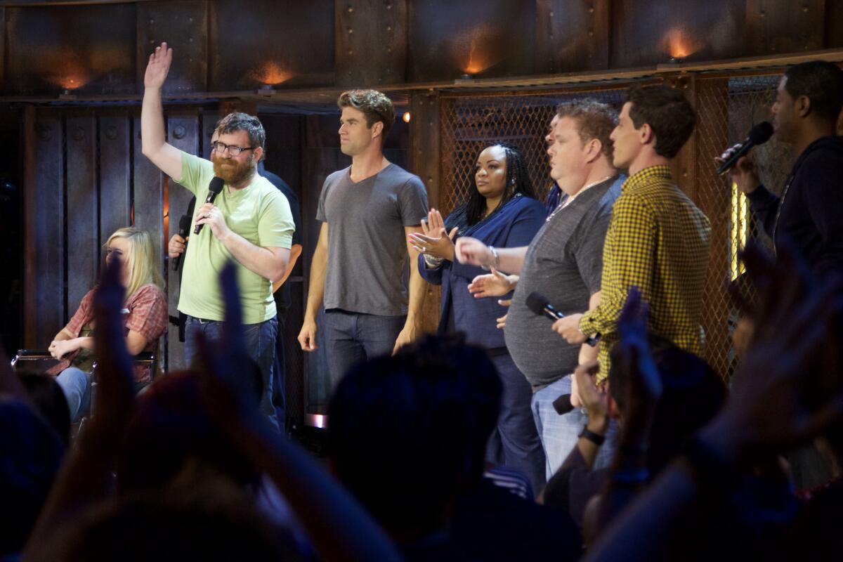 About half a dozen people stand on a small stage. A bearded man in a green T-shirt holds a microphone and waves.