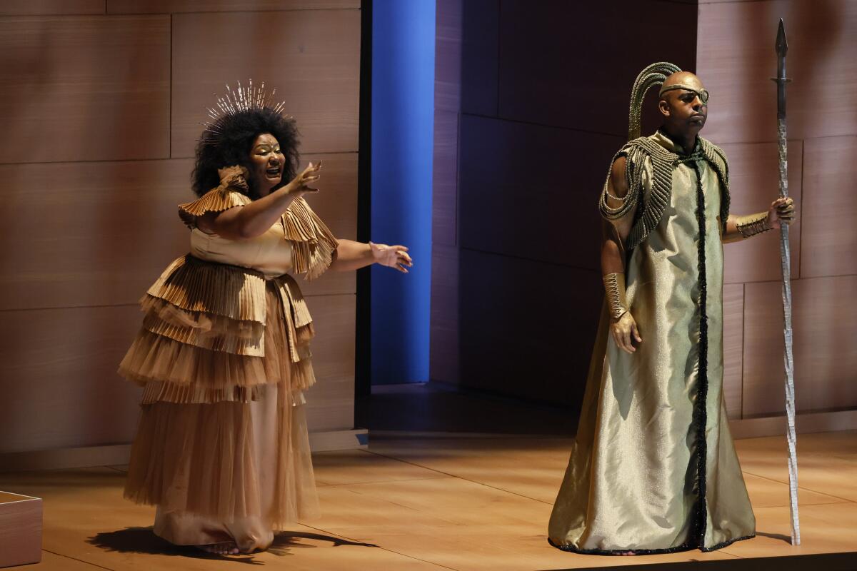 A woman in an elaborate shimmering skirt motions to her husband in dashing tribal dress in "Das Rheingold" at Disney Hall.
